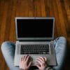 Man sitting cross-legged on hardwood floor, hands on laptop typing