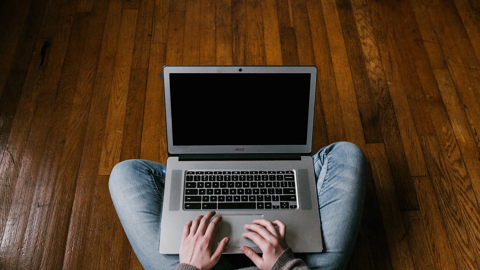 Man sitting cross-legged on hardwood floor, hands on laptop typing