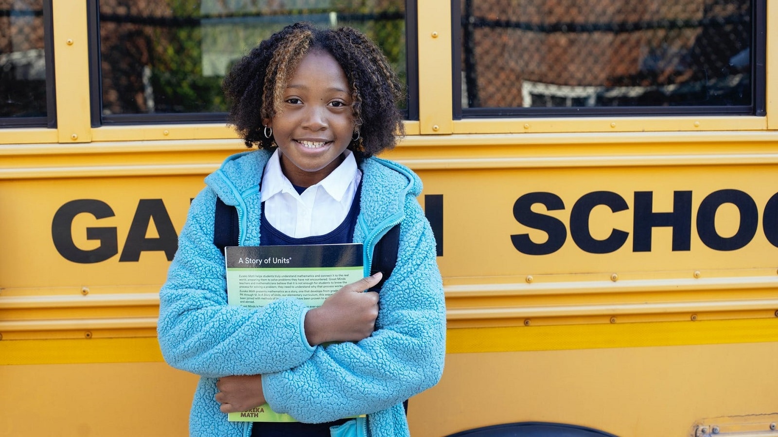 Schoolgirl hugging textbook in front of yellow school bus