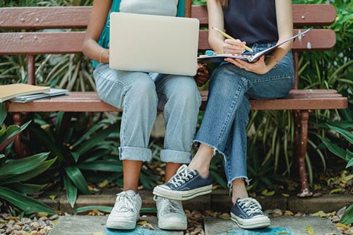 Two people sitting on a bench writing notes while looking at a laptop