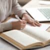 Closeup photo of a female student writing down notes with a textbook and laptop open on her desk
