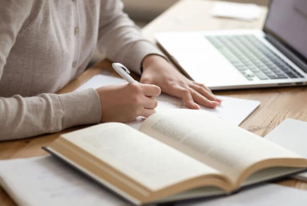Closeup photo of a female student writing down notes with a textbook and laptop open on her desk