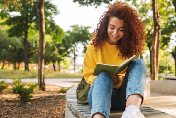 Young curly haired student girl sitting outdoors in nature park writing notes in notebook.