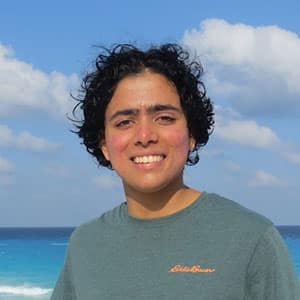 A smiling student with wind blown hair standing on the back with the water and blue sky in the background.