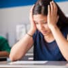 Hispanic teen with head between her hands looking worried staring down at a sheet of paper on a desk with students behind her writing at their desks