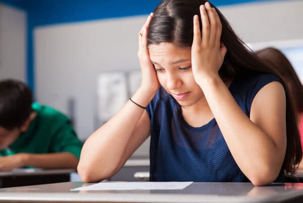 Hispanic teen with head between her hands looking worried staring down at a sheet of paper on a desk with students behind her writing at their desks