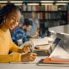 A high school student in a library setting writing notes on paper with a laptop in front of her with other students and book shelves in the background