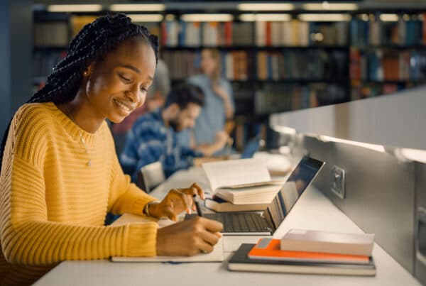 A high school student in a library setting writing notes on paper with a laptop in front of her with other students and book shelves in the background