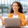 Young female student with eyes closed smiling and hands up level with her head and laptop in front of her