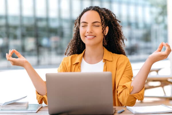 Young female student with eyes closed smiling and hands up level with her head and laptop in front of her