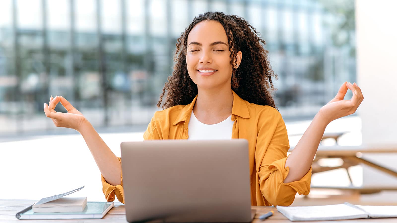 Young female student with eyes closed smiling and hands up level with her head and laptop in front of her