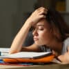 Tired student with her head resting in her hand with a stack of folders and notebooks on her desk and a cup of coffee nearby
