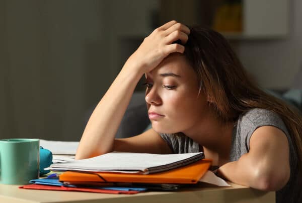 Tired student with her head resting in her hand with a stack of folders and notebooks on her desk and a cup of coffee nearby