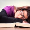 Student wearing glasses with head down sleeping on a book with a stack of books around her