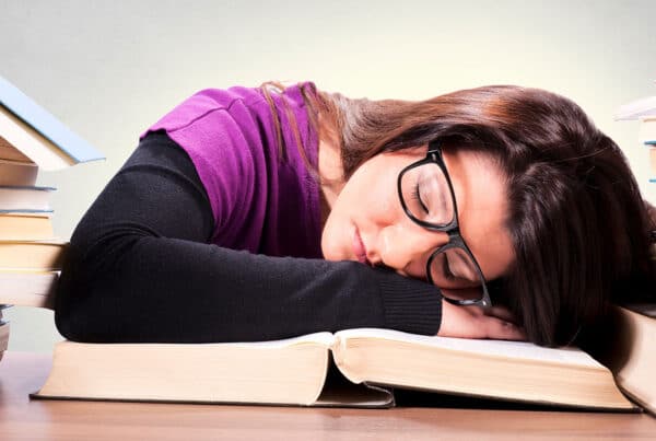 Student wearing glasses with head down sleeping on a book with a stack of books around her