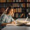 Female student in a library sitting at a desk reading journals and books studying with a soft smile on her face