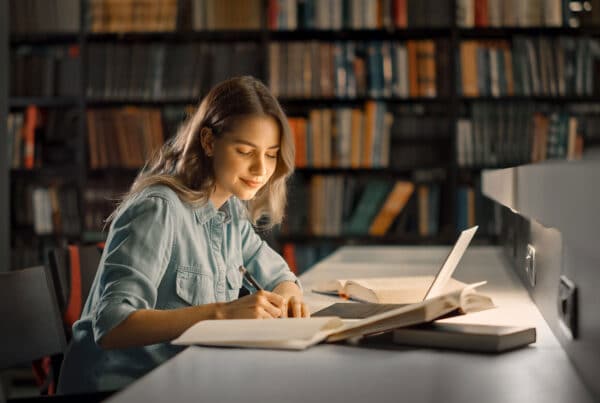 Female student in a library sitting at a desk reading journals and books studying with a soft smile on her face