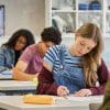 Image of several students in a row of desks with a focus on a female student looking down as she writes on a test she and others in the room are taking