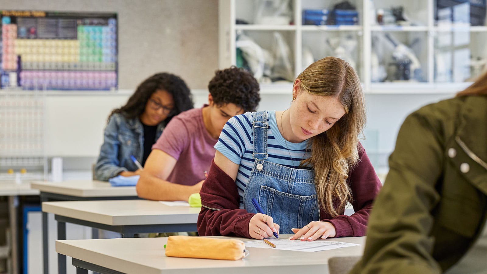 Image of several students in a row of desks with a focus on a female student looking down as she writes on a test she and others in the room are taking
