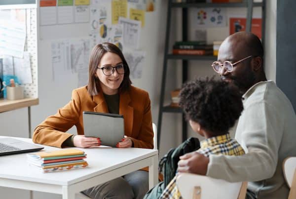 Image of a teacher behind a desk talking to a father and son seated next to her discussing the students academic progress