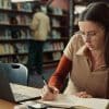 Image of a girl with long dark hair at a desk in a library writing while looking at a laptop