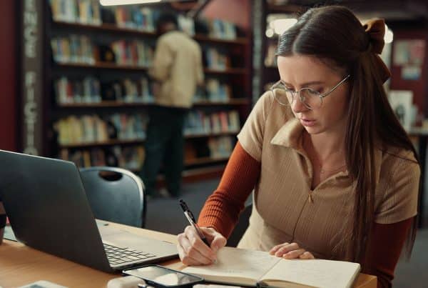 Image of a girl with long dark hair at a desk in a library writing while looking at a laptop