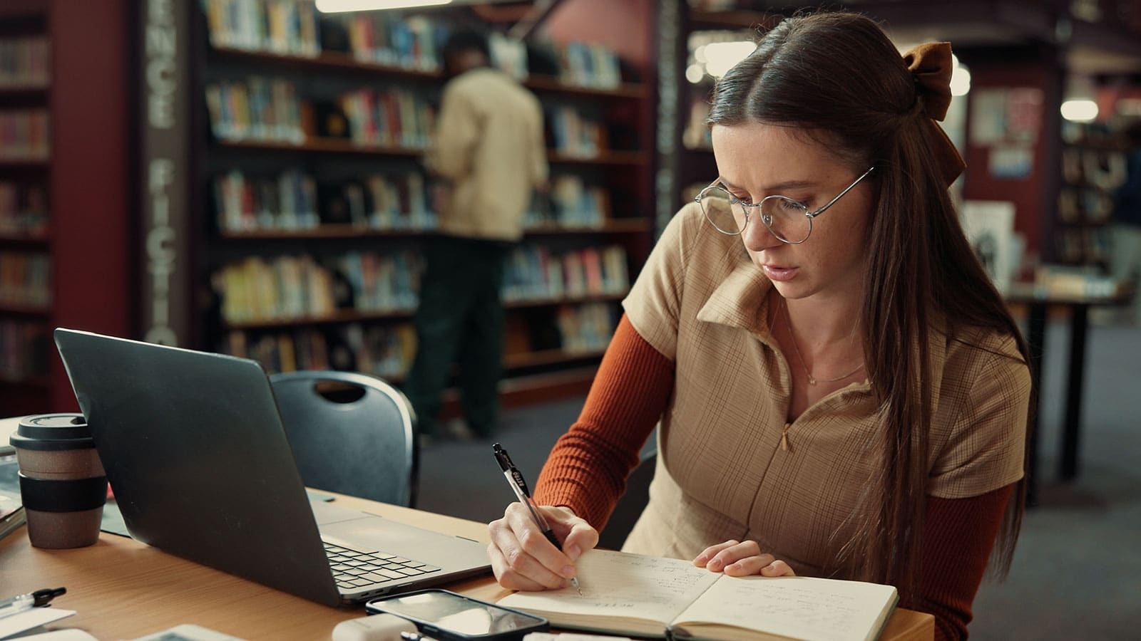 Image of a girl with long dark hair at a desk in a library writing while looking at a laptop