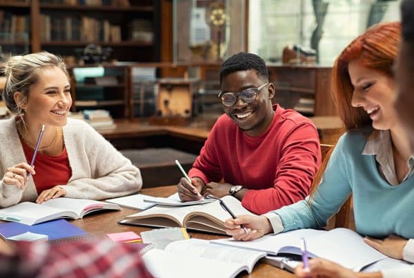 Image of several college students sitting at a table in a library smiling with open books in front of them as they study