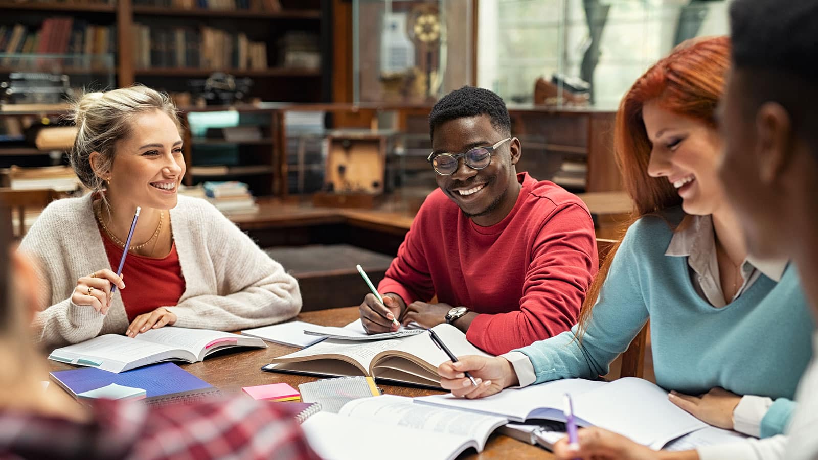 Image of several college students sitting at a table in a library smiling with open books in front of them as they study