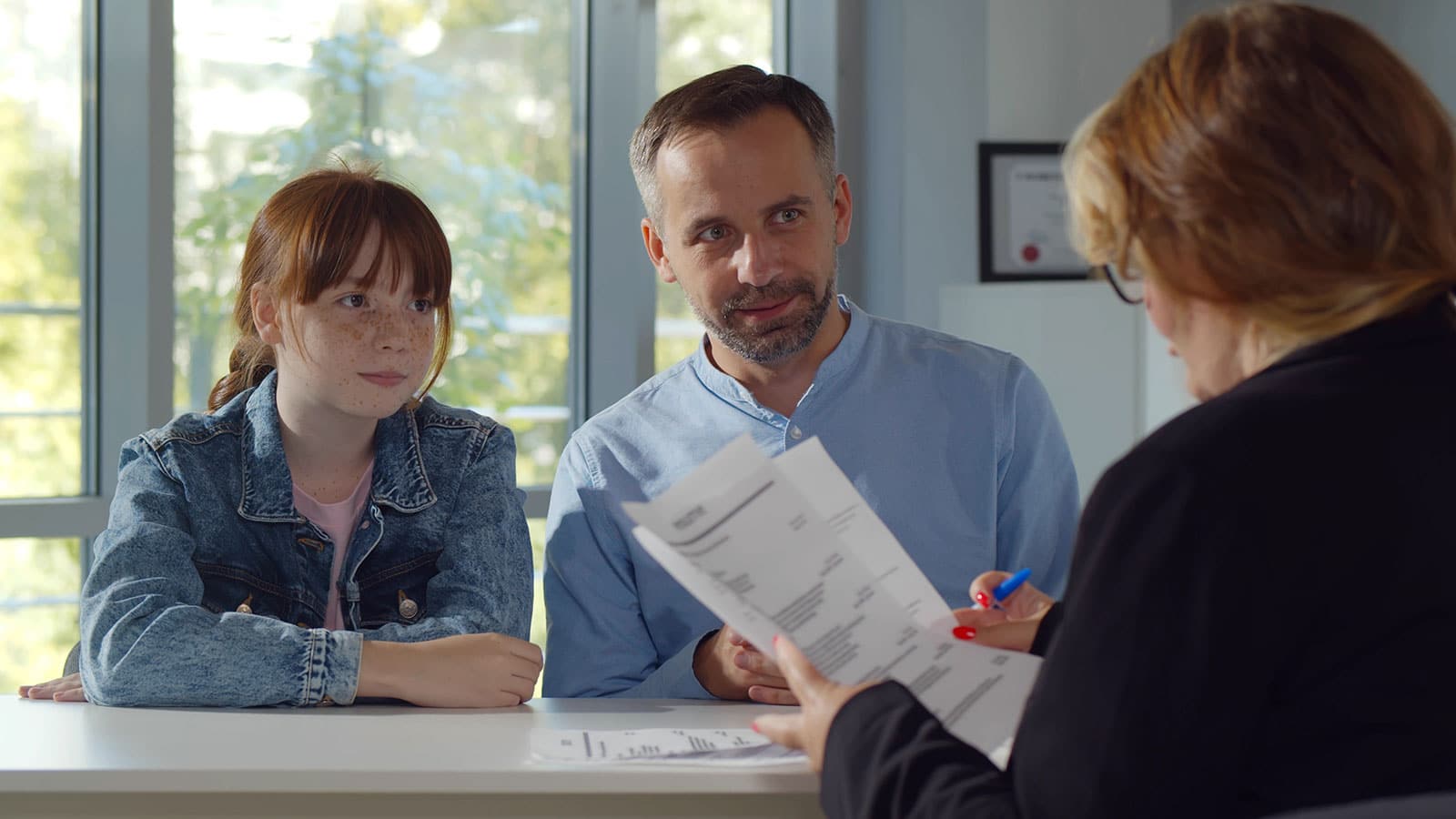 Image of a father and daughter sitting behind a desk with a back view of a woman looking over paperwork