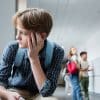 A child wearing a back pack sitting alone staring out the window with a school hallway and other students socializing in the background