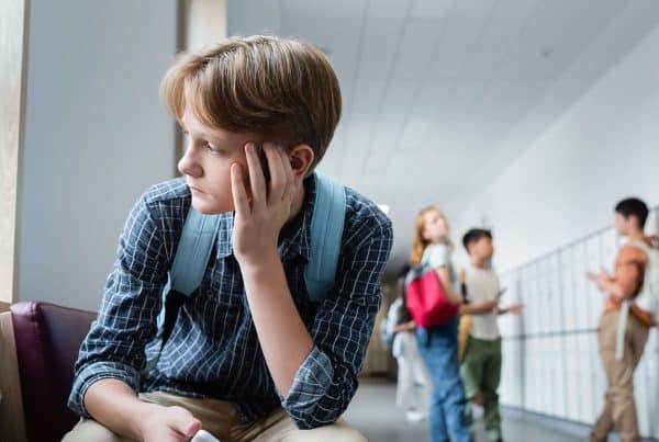 A child wearing a back pack sitting alone staring out the window with a school hallway and other students socializing in the background
