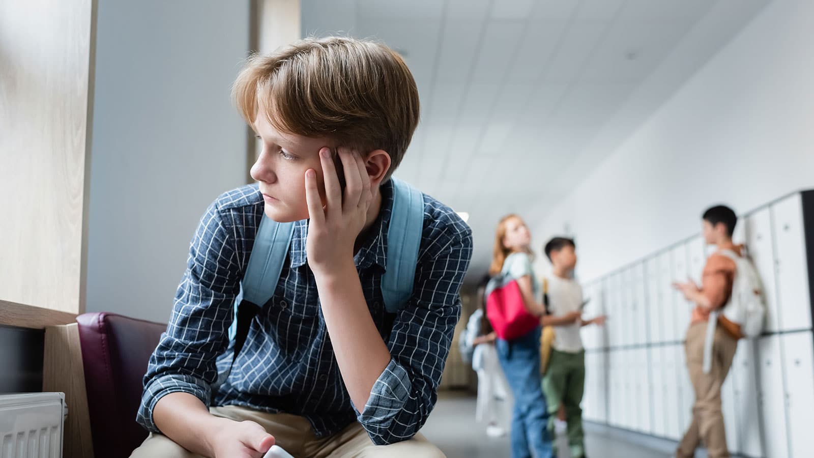 A child wearing a back pack sitting alone staring out the window with a school hallway and other students socializing in the background