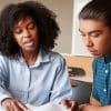 An instructor next to a student with her hand on a desk showing the student something on a page